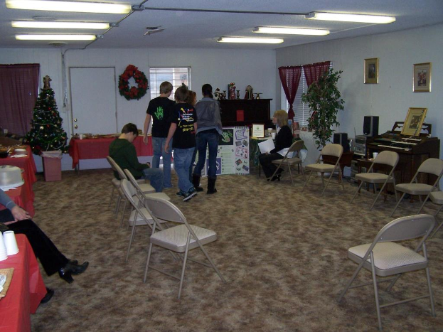 Ein Raum mit mehreren Stühlen, Tischen und Personen, einige stehen, einige sitzen, mit Tassen und Behältern auf den Tischen, einem Weihnachtsbaum, einem Piano auf der rechten Seite, Foto-Rahmen, Fenstern und Rohrleuchten an der Decke sowie einer Wand mit einem Strauß, Vorhang, Tür und weiteren Foto-Rahmen im Hintergrund.