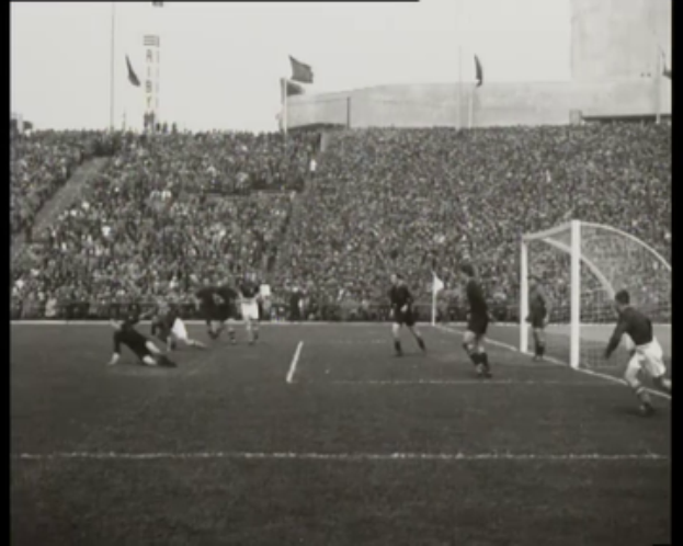 Ein Schwarz-Weiß-Foto eines WM-Finales 1958 zwischen Manchester United und Liverpool mit Spielern auf dem Feld, einem Torpfosten rechts, Zuschauern in den Rängen und Fahnen im Hintergrund.