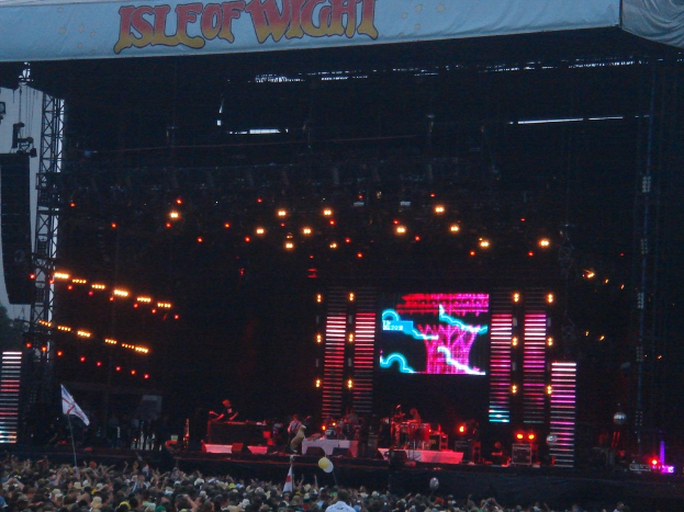 Eine Bühne mit Lichtern, einem Bildschirm und Musikern mit Instrumenten, mit einem Banner oben, einer Menge unten und Fahnen mit Stangen und Ballons drumherum.