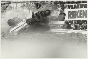 Ein Schwarz-Weiß-Foto eines demolierten Rennwagens umgeben von Reifen, mit einer Gruppe von Menschen rechts und einer Tafel mit Text im Hintergrund.