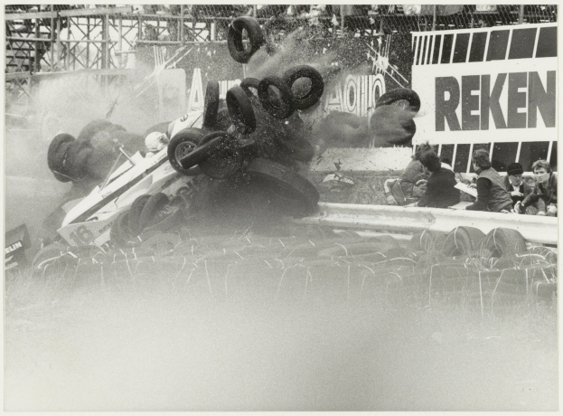 Ein Schwarz-Weiß-Foto eines demolierten Rennwagens umgeben von Reifen, mit einer Gruppe von Menschen rechts und einer Tafel mit Text im Hintergrund.