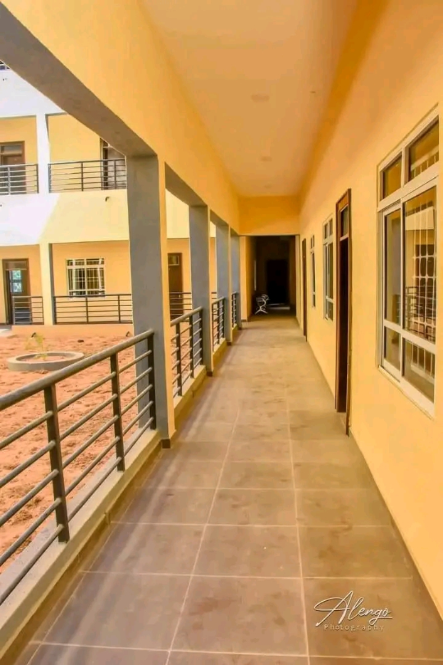 Ein Gebäude mit Fenstern, Türen, Säulen, Geländern und einem langen Flur mit einem gefliesten Boden und einem Balkon, mit anderen Gebäuden und einem klaren blauen Himmel im Hintergrund, und einem Text unten, der "3-Zimmer-Wohnung zur Miete in Ntinda, Kampala" lautet.