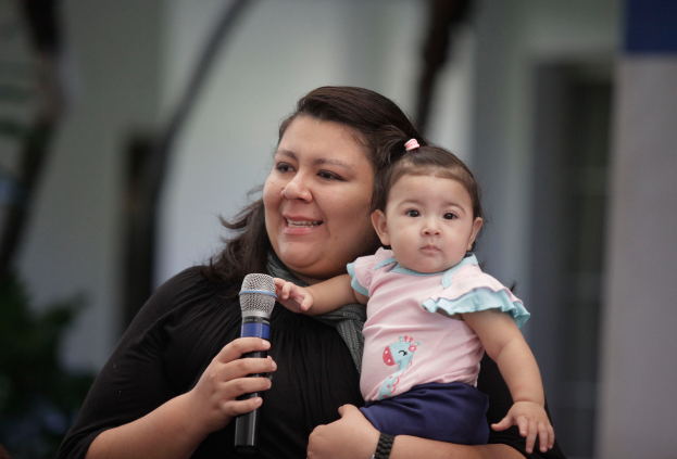 Eine Frau in einem weißen Hemd und blauen Jeans hält ein Baby im Arm und spricht in ein Mikrofon mit einem warmen Ausdruck, während das Baby sie mit einem zufriedenen Lächeln ansieht.