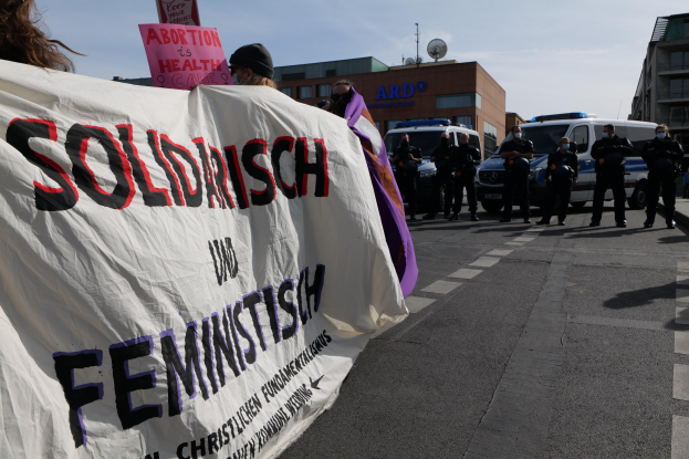 Gruppe von Menschen marschiert die Straße entlang und hält ein 'Solidarität und Feminismus'-Schild, mit parkenden Fahrzeugen, Gebäuden, einer Schüsselantenne und einem klaren blauen Himmel im Hintergrund.