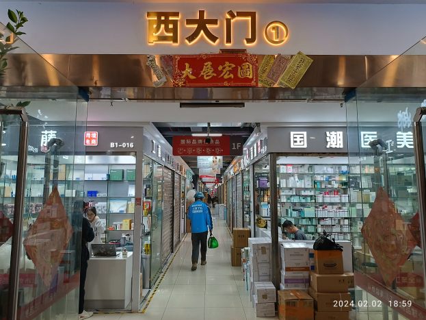 Innenraum einer chinesischen Apotheke mit Glaswänden, Regalen voller Gegenstände, verstreuten Kartons, einer Pflanze auf der linken Seite und einer Namensschild oben.