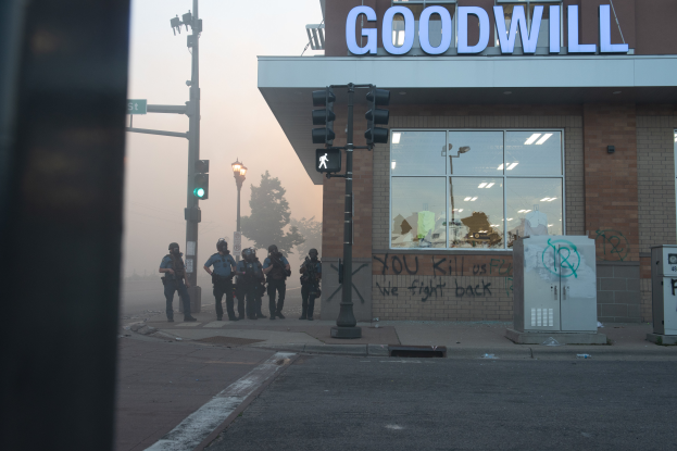 Eine Gruppe von Polizisten in Helmen und mit Waffen in der Hand steht vor einem Goodwill-Laden mit Glaswänden, mit Laternenmasten, Verkehrsampeln, Bäumen und einem klaren Himmel im Hintergrund.
