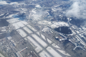 Luftaufnahme eines Flughafens mit schneebedecktem Boden, Gebäuden, Straßen, Brücken, Wasser, Bäumen und Wolken am Himmel.