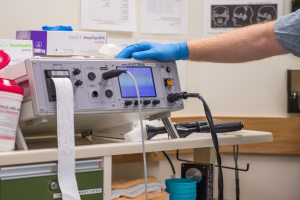 Eine Person in blauen Handschuhen arbeitet an einem medizinischen Gerät auf einem Tisch, umgeben von verschiedenen Gegenständen, mit Papieren an der Wand in einer Krankenhausumgebung.