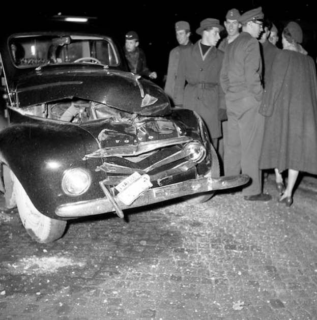 Eine Schwarz-Weiß-Aufnahme eines beschädigten Autos am Straßenrand, umgeben von einer Gruppe besorgter Menschen.