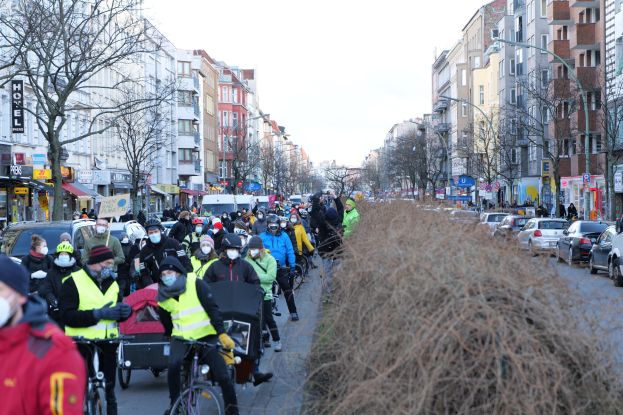 Eine große Gruppe von Menschen in Masken und Sicherheitswesten, die auf Fahrrädern eine von Bäumen gesäumte Straße entlangfahren, mit Gebäuden, Laternenmasten, Texttafeln, Fahrzeugen, trockenem Gras und einem klaren blauen Himmel.