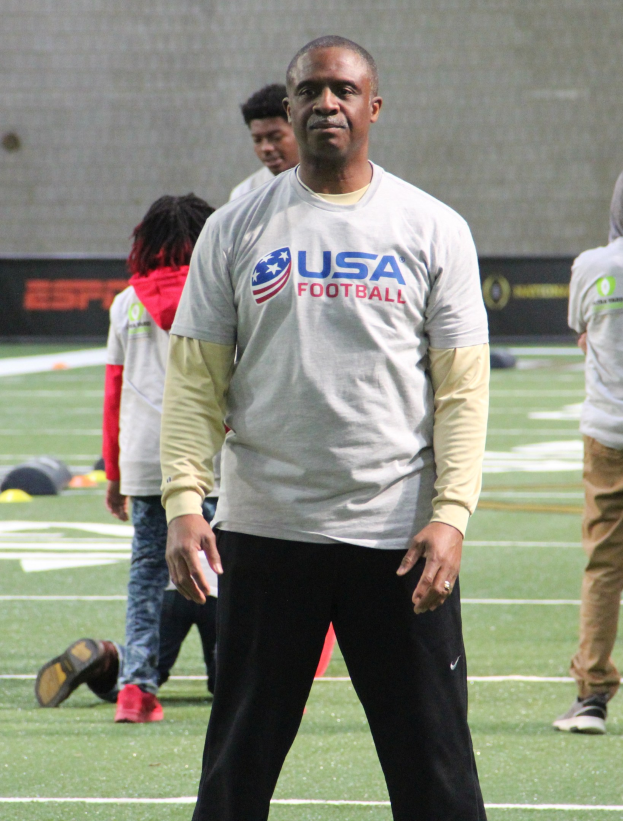 Ein Fußballspieler in Fußballschuhen steht auf einem Feld neben einer Gruppe von Menschen und einer Wand im Hintergrund.