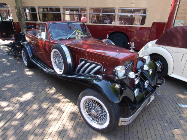 Ein Oldtimer neben einem Doppeldeckerbus auf Kopfsteinpflasterstraße, mit Stühlen und Baum im Vordergrund, einem Gebäude im Hintergrund und Menschen im Bus.