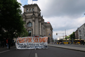 Eine Gruppe von Menschen marschiert eine baumbestandene Straße entlang und hält ein Banner mit der Aufschrift "Kampf für Solidarität", mit Gebäuden, Fahrzeugen und einer Flagge auf einem Gebäude im Hintergrund.