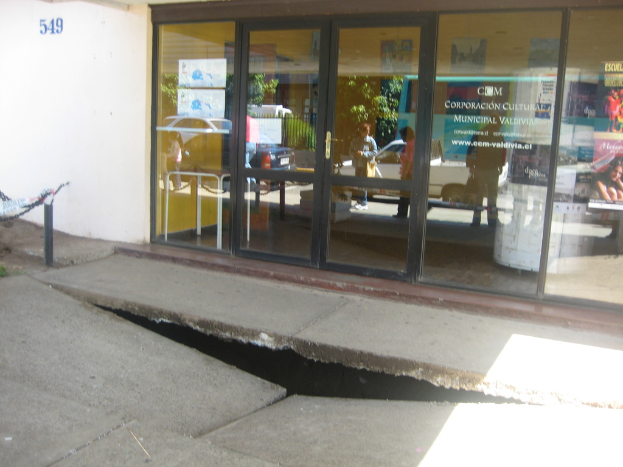 Eine gebrochene Oberfläche mit einer Glastür, Wand und Pfahl im Hintergrund; die Spiegelung zeigt fahrende Fahrzeuge und einige stehende Personen.