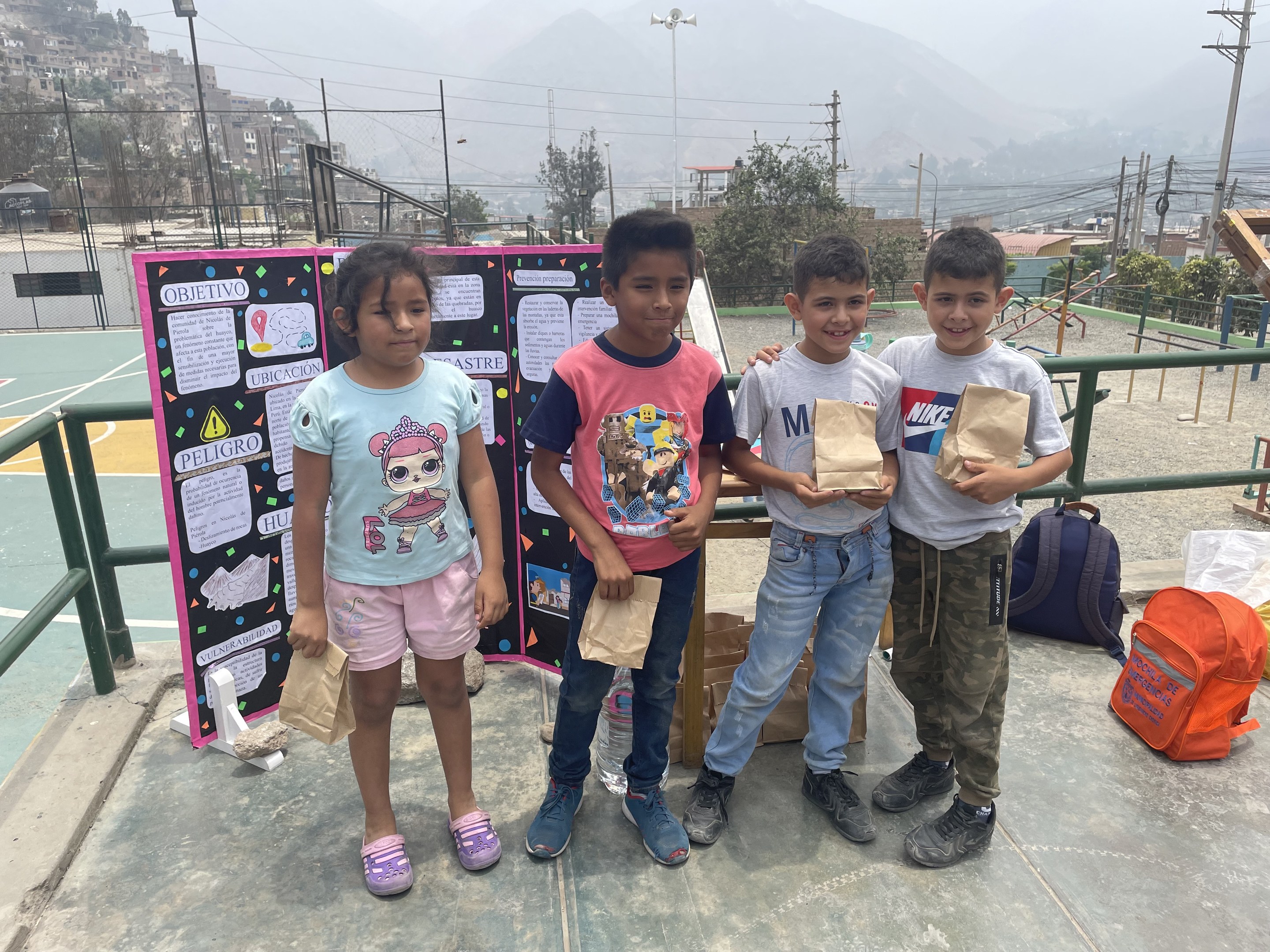 Gruppe von Kindern auf einem Basketballfeld mit Papiertüten in den Händen, mit Gegenständen auf der rechten Seite und einer Tafel mit Text auf der linken Seite, vor einem Hintergrund aus Zaun, Pfosten, Straßenlaternen, Bäumen, Gebäuden, Hügeln und Himmel.