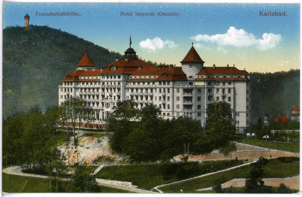 Eine alte Postkarte, die das Hotel Imperial in Karlsbad, Deutschland zeigt, ein großes Gebäude mit vielen Fenstern, umgeben von Bäumen, Pflanzen und Gras, mit einem Weg, einer Baumgruppe, Hügeln und einem bewölkten Himmel im Hintergrund und Text oben.