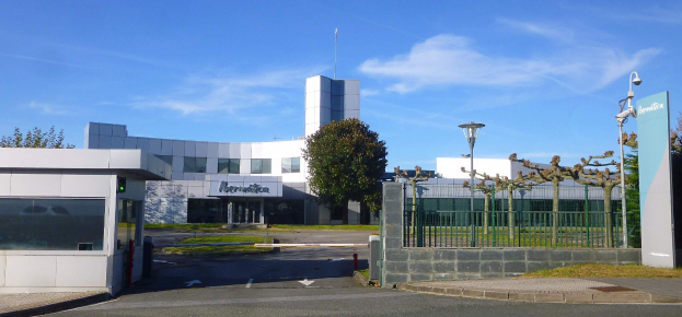 Ein großes Gebäude mit zahlreichen Fenstern und einem Tor davor, umgeben von Straßenmasten, Laternen, Gittern, Bäumen und Gras, unter einem bewölkten Himmel, das als Hauptsitz von Iberostica in den Niederlanden dient.