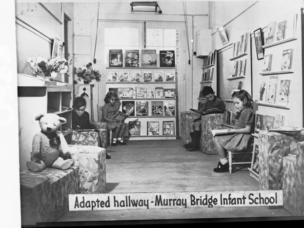 Eine Gruppe von Kindern sitzt auf Stühlen in einem Raum mit Bücherregalen, einem Teddybär, einer Blumenvase auf dem Tisch und gerahmten Gegenständen an der Wand, mit dem Text "Angepasster Flur Murray Bridge Kindergarten" unten.