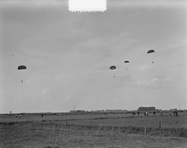 Ein Schwarz-Weiß-Foto einer Gruppe von Menschen beim Segelflug, mit offenen Fallschirmen und Bäumen und Gebäuden im Hintergrund, mit Gras am Boden.