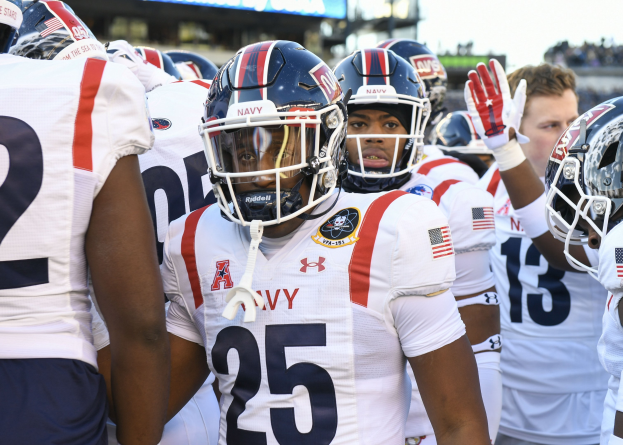 Gruppe von Männern in Fußballausrüstung auf einem Feld mit Helmen und Handschuhen, Hintergrund zeigt unscharfe Zuschauer, ein Scoreboard mit 'Auburn vs Virginia Tech - 11/28/15 College Football Pick, Odds, and Prediction' und Himmel.