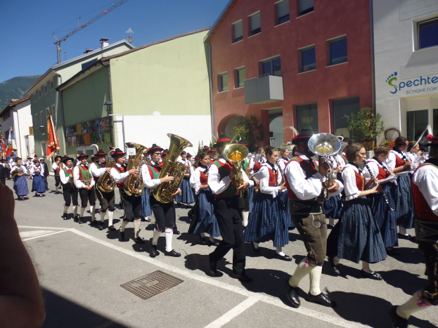 Eine Gruppe von Menschen in traditioneller bayrischer Tracht, die auf der Straße spielen, während sie durch eine Straße mit Gebäuden gehen, einige halten Fahnen, mit einem Hügel und einem klaren blauen Himmel im Hintergrund.