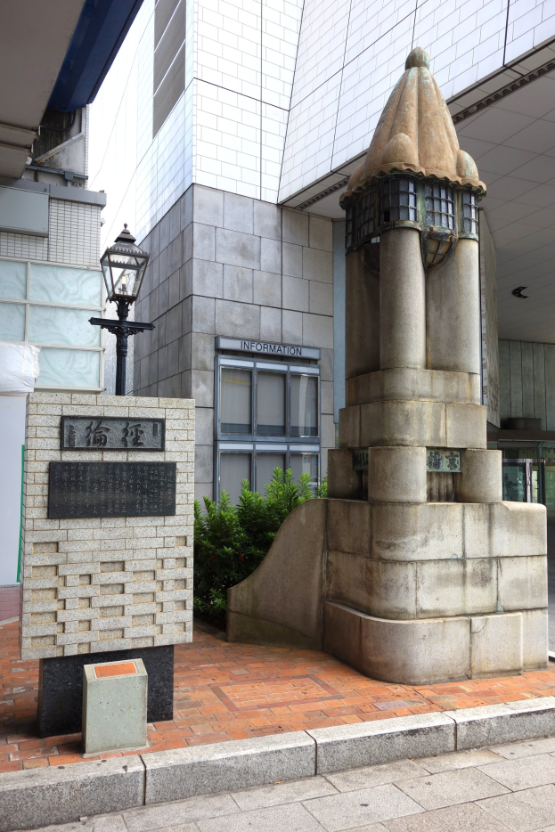 Ein Denkmal steht in der Mitte einer Stadtstraße, umgeben von Gebäuden, Straßenmöbeln und Grün unter einem klaren blauen Himmel.