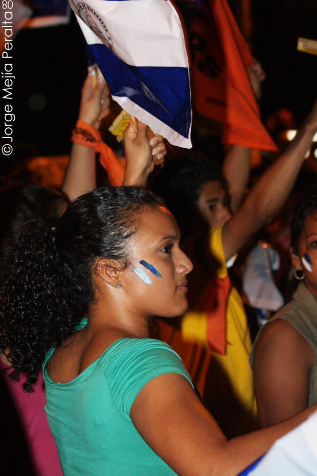 Eine Frau in einem grünen T-Shirt steht bei einer Demonstration und hält ein weißes Schild in der Hand, während viele andere Menschen hinter ihr stehen.