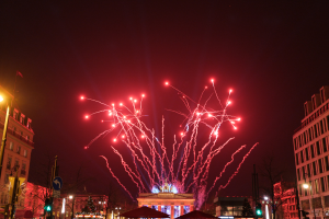 Eine Stadtstraße in Berlin an Silvester, voller Gebäude, Bäume, Laternenpfähle, Verkehrszeichen, Zelte und Menschen, mit einem von Feuerwerk erhellten Himmel im Hintergrund.