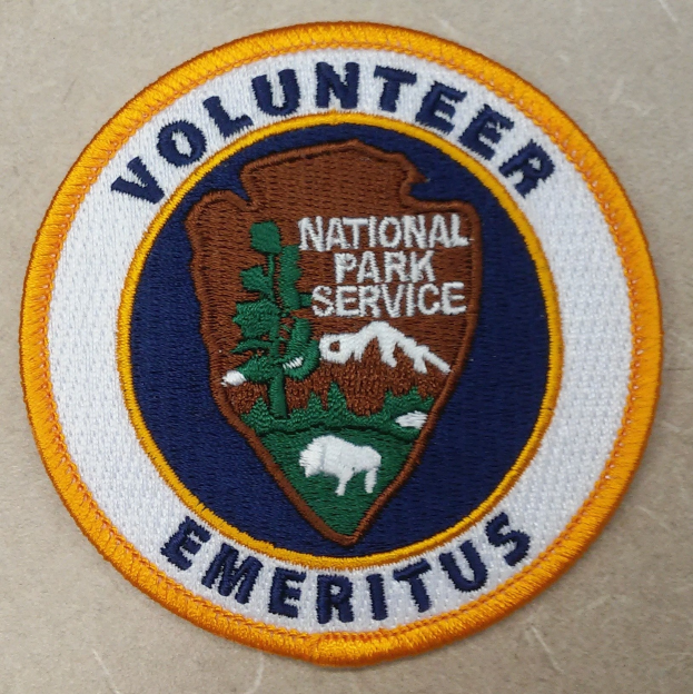 Runder Aufnäher mit "Volunteer National Park Service Emeritus" in fetter schwarzer Schrift auf weißem Hintergrund, umrandet in blau mit einem gelben Kreis um den Text.
