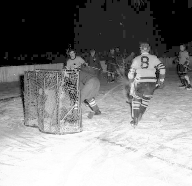 Schwarzes und weißes Foto von Männern, die auf einem Eisplatz Hockey spielen, mit einem Netz im Vordergrund und einer Wand im Hintergrund.