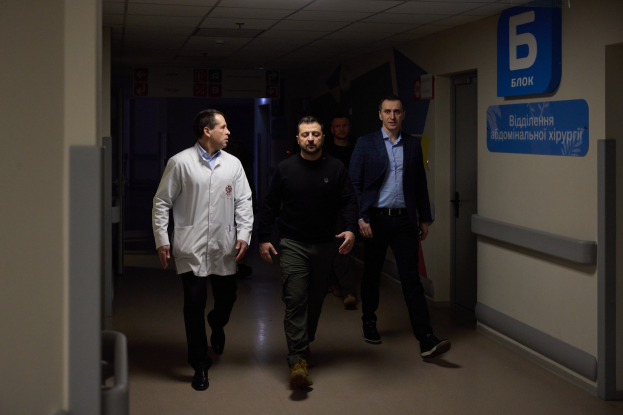 Eine Gruppe von Männern geht einen hell erleuchteten Krankenhausflur entlang und sieht besorgt aus, mit einer Tür auf der rechten Seite und Texttafeln auf der linken Seite.