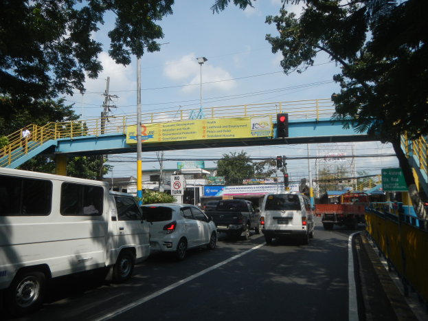 Eine belebte Straße mit Autos und Lastwagen, eine Brücke mit Geländern und Treppen, Laternenmäste, Verkehrsampeln, Schilder mit Text, Bäume, Gebäude und ein bewölkter Himmel.