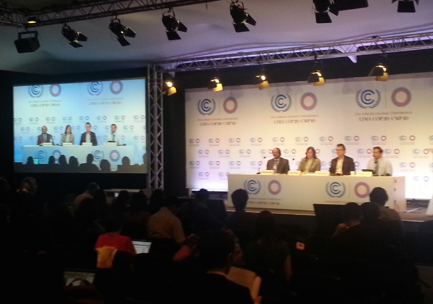 Menschen sitzen auf Stühlen auf einer Bühne mit Namensschildern und Mikrofonen auf einem Tisch davor, während Zuschauer in Stühlen sitzen und zuschauen; ein Projektionsschirm und Beleuchtung an Eisenstangen sind im Hintergrund sichtbar.