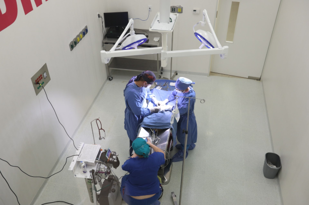 Chirurgen führen einen Eingriff an einem Patienten in einem Operationssaal durch, mit medizinischer Ausrüstung an einer Wand und einem Objekt auf dem Boden rechts.