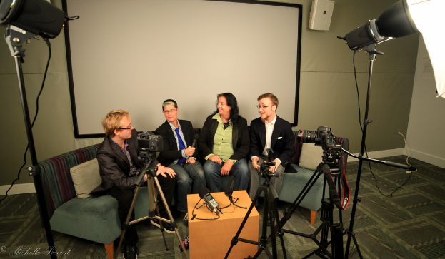 Eine Gruppe von drei Personen auf einem Sofa sitzend, wobei eine ein Mikrofon hält, vor einem Projektionsschirm interviewt, mit Kameras und Beleuchtungsausrüstung drumherum.