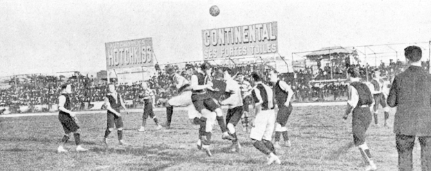Ein Schwarz-Weiß-Bild einer Gruppe von Männern, die auf einem Feld Fußball spielen, mit einem Ball in der Luft und Zuschauern im Hintergrund, mit Schildern, die Text über das Spiel anzeigen.