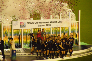Eine Gruppe von Fussballspielerinnen in schwarzen Uniformen feiert ihren Sieg auf einer Bühne, umgeben von einer Menge mit Kameras, mit einem dekorierten Hintergrund und einem grasbewachsenen Boden.