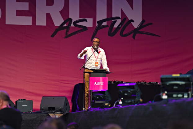 Mann in weißem Hemd und braunen Hosen, der in ein Mikrofon spricht, vor einem pinken 'Berlin as Fuck'-Schild und einer Menge