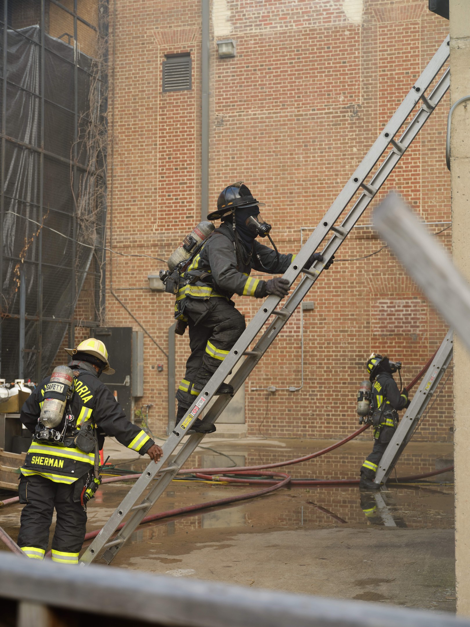 Feuerwehrleute in Helmen und Zylindern klettern an einer Leiter vor einem Backsteingebäude mit Rohren auf dem Boden und einer Metallstange am Boden, mit einem anderen Gebäude und einem Netz im Hintergrund.
