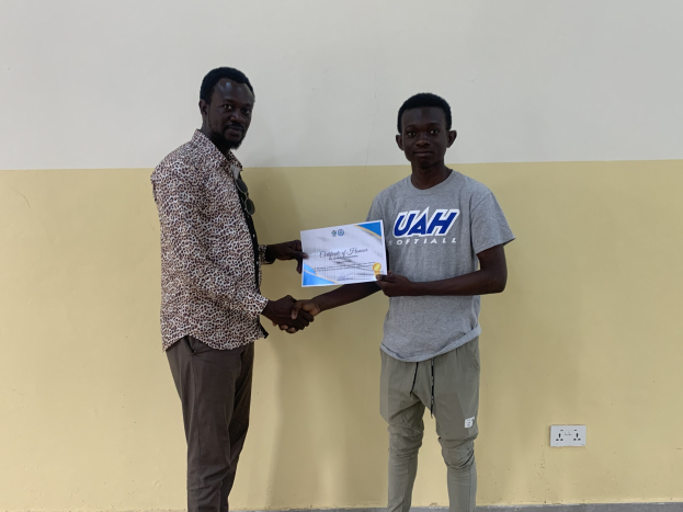 Zwei Männer, die sich vor einer gelben Wand die Hand schütteln, wobei der Mann links ein Zertifikat hält, nachdem er ein Stipendium von der Universität Ghana erhalten hat.