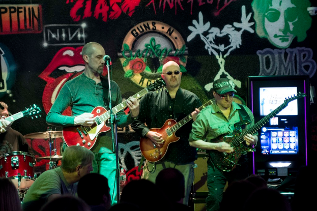 Drei M├Ąnner, die auf einer Buhne Gitarren spielen, mit einer Menge darunter und einem Graffiti oder einer Wandmalerei im Hintergrund.