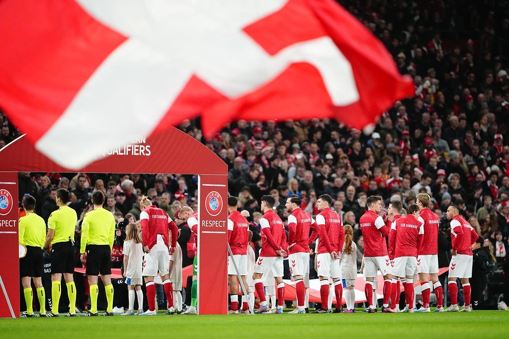 Gruppe von Menschen auf einem Fußballfeld mit einer roten und weißen Flagge im Vordergrund, einem Bogen mit der Aufschrift "Bayern Munich vs Bayern Munich Betting Tips & Preview" im Hintergrund und einer großen Menge im Stadion.