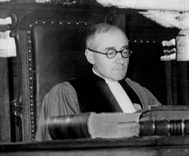 Ein Schwarz-Weiß-Foto eines Mannes mit Brille und Robe, der an einem Schreibtisch mit einem Buch sitzt und ernst dreinschaut.