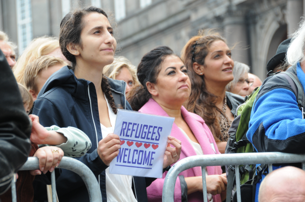 Eine Gruppe von Menschen vor einem Gebäude stehend, mit einer Frau in der Mitte, die ein "Flüchtlinge Willkommen"-Schild hält, andere mit Taschen, und Geländer im Vordergrund.