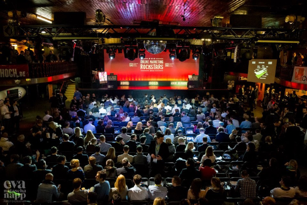 Eine große Menge Menschen sitzt in Reihen vor einer hell erleuchteten Bühne mit Bildschirmen und Bannern, was auf ein Event wie eine Online-Marketing-Konferenz hindeutet.