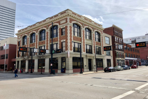 Großes Backsteingebäude an einer Straßenkreuzung mit umliegenden städtischen Strukturen, Fahrzeugen, Fußgängern und Bäumen unter einem bewölkten Himmel.