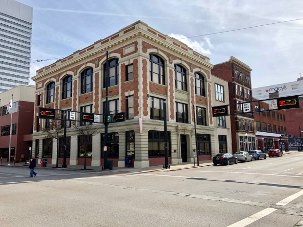 Großes Backsteingebäude an einer Straßenkreuzung mit umliegenden städtischen Strukturen, Fahrzeugen, Fußgängern und Bäumen unter einem bewölkten Himmel.