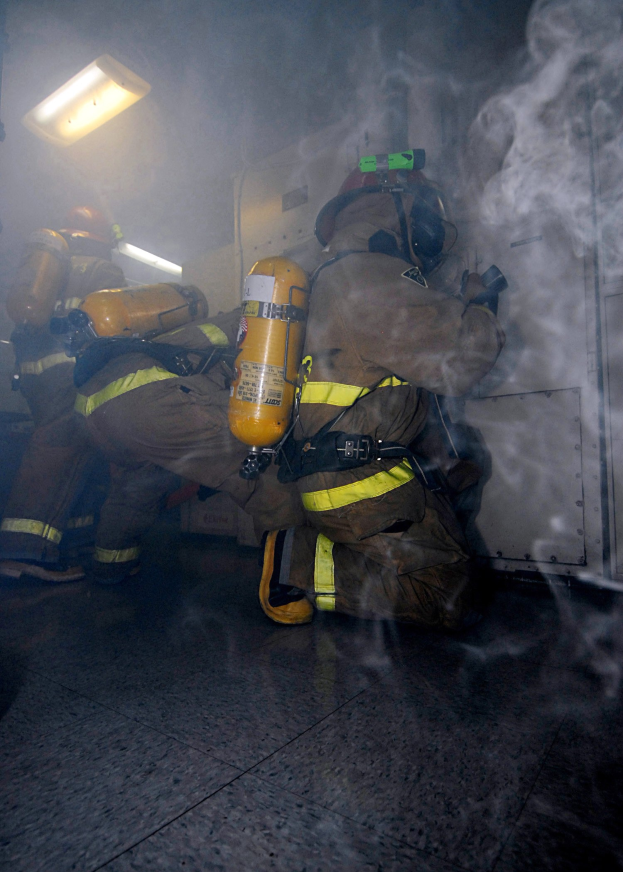 Feuerwehrleute in einem raucherfüllten Raum mit beleuchteter Ausrüstung und einer Wand im Hintergrund.