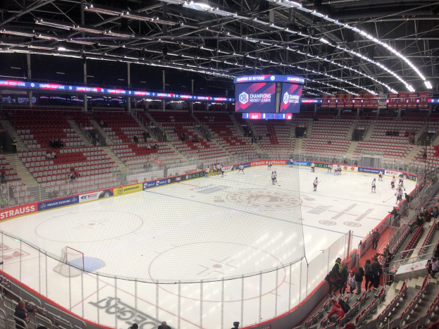 Eishockeyspiel in einer großen Arena zwischen den St. Louis Blues und Detroit Red Wings, mit Zuschauern auf Sitzen und Stehplätzen in der Nähe der Eisfläche umgeben von einem Zaun und Metallrahmen, unter einer Decke mit Deckenleuchten und einem Anzeigebildschirm.