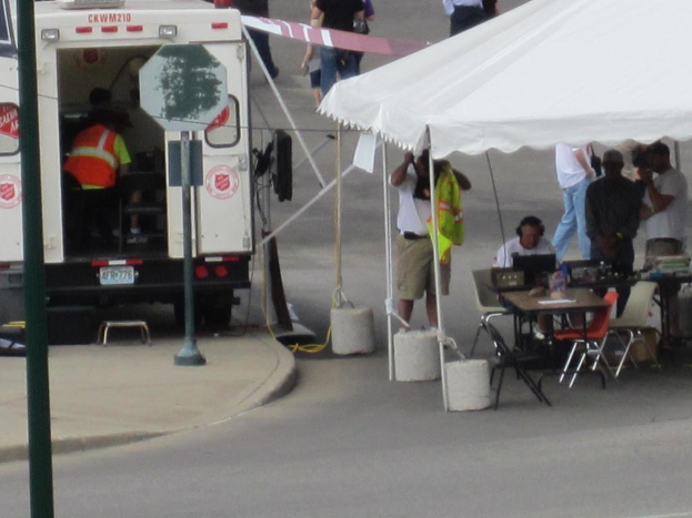 Ein Rettungswagen steht auf der linken Seite einer Stra├če, waehrend auf der rechten Seite ein Zelt viele Menschen sch├╝tzt, mit einem Tisch und einem Stuhl in der N├Ąhe.